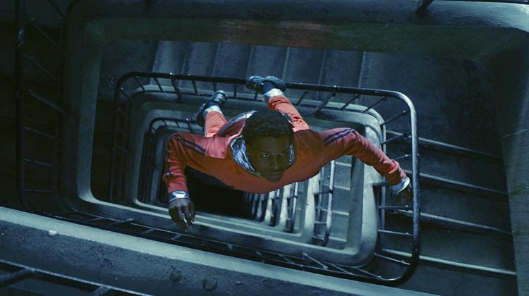 A rectangular stair well vanishes into darkness and floating in the middle of the space is a youth dressed in clothing reminiscent of an astronaut  