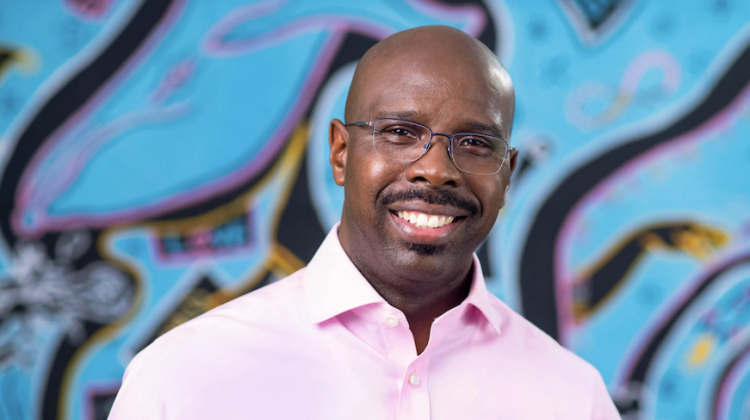 Headshot of Mike Johnson wearing a pink shirt against a blue, pink, black and yellow art background