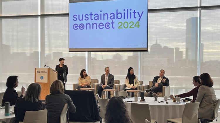 One person stands behind a lectern with four seated panelists to her left. Above them, a screen displays &quot;Sustainability connect 2024. Boston’s skyline fills the windows behind them. 