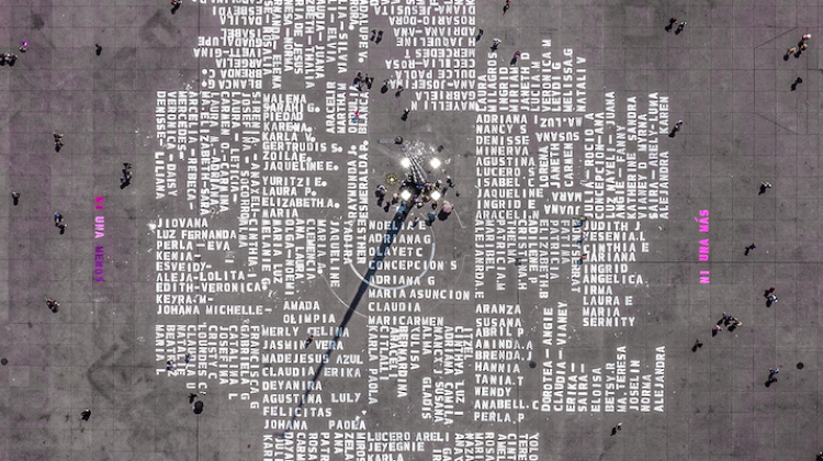 White names are written in blocks, surrounding a lighted pillar with offerings and remembrances. The names are larger than people and their layout mirrors the concrete tiles that make up the plaza. At the four compass points, in pink. is written "ni una más" not one more. 