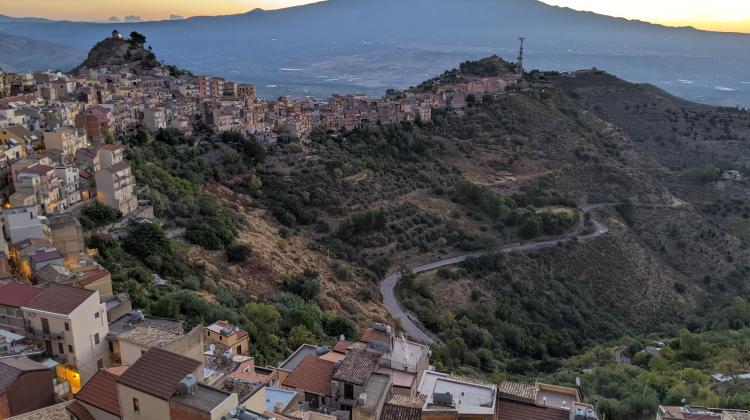 Landscape of a village perched atop the ridge line of a mountain range. The village follows the ridge line from below the image, across the left edge, and into the middle of the photo. In the background another, distant mountain range is visible.