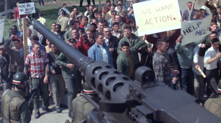 In the background of this image is a group of individuals carrying signs that appear to be protestors, in the foreground is the barrel and magazine of a machine gun overlooking the crowd.