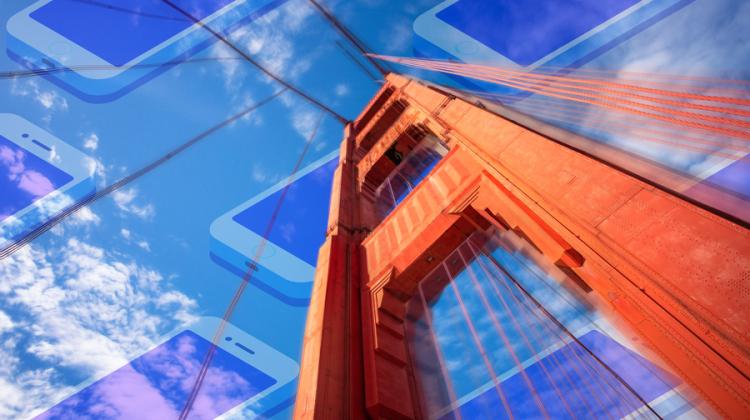 Image of a central support arch for the Golden Gate bridge, taken from the base looking upwards. The blue, semi-cloudy sky has digital devices mapped across, semi-transparently. 