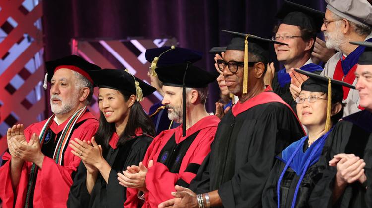 Eight men and women in academic regalia applauding on a stage indoors