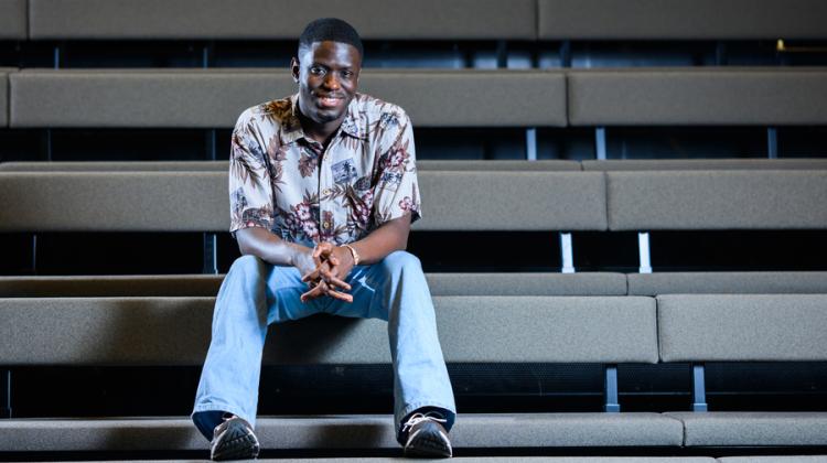 Kwesi Afrifa sits on the bleachers of a theatre with hands clasped. The rows of bleachers create strong horizontal lines.