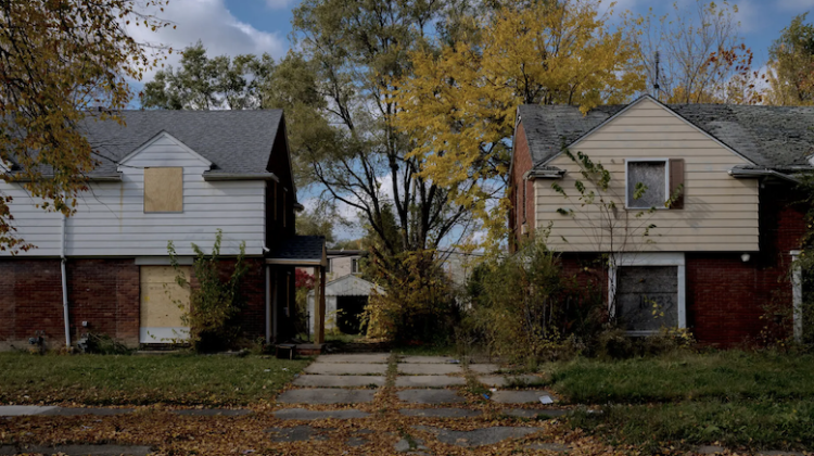 Two houses with the windows and doors boarded up sit next to each other amidst overgrown shrubs.