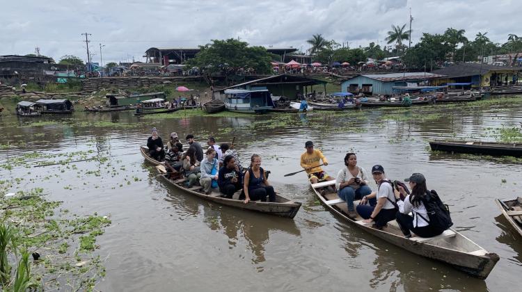 Image of three boats, lined with students. In the background a small town and market can be seen.