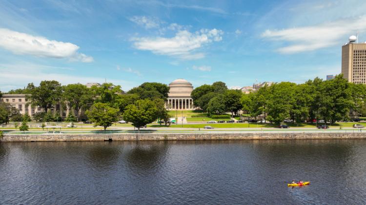 Drone photo shows the Charles River and MIT campus, with Killian Court in the middle, on a sunny day. Two people are in a kayak.