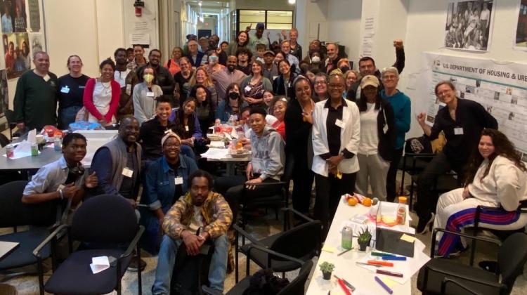 A diverse group of folks gathered in a room in building 7 at MIT, images of various posters/plots are hung on walls and desks and tables indicate where folks have been working together