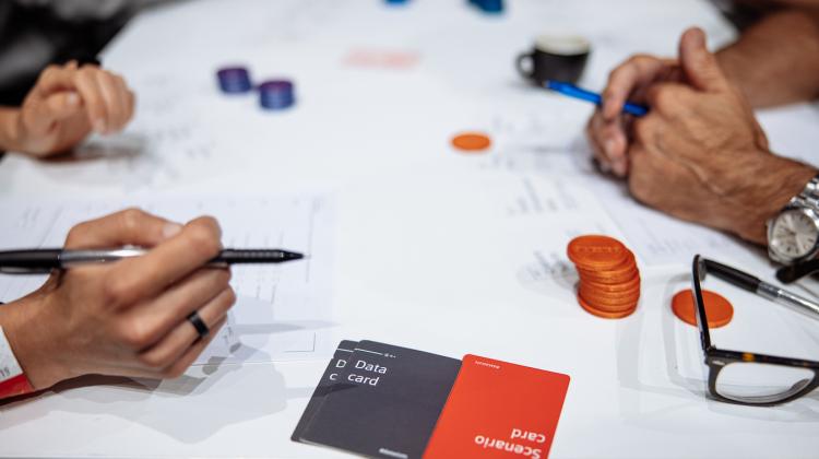 Shot of multiple individuals playing Data Slots in-person. The image centers on their hands, holding pens with game pieces and cards on the table.