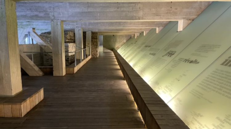The Abolition of Slavery Memorial Museum in Nantes, a long walkway is defined by columns on the left and angled glass with text on the right. The space is underground and lit by indirect lighting.
