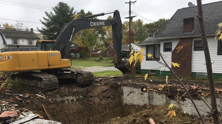 Image of a home demolition in Youngstown Ohio. On the left a large digger is removing debris. Next to the digger a hole can be seen that one understands to be a basement of the demolished home. In the background, other single family homes are visible.