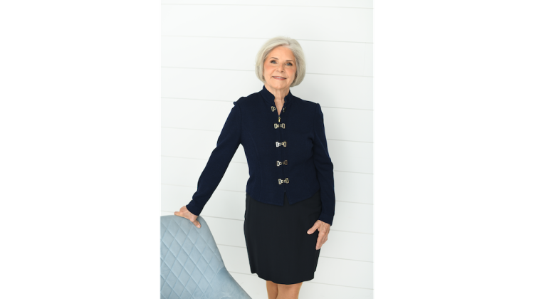 Photo of Amy Glasmeier, founder of the MIT Living Wage Calculator, posing in a blue suit next to a chair.