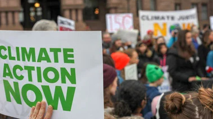 An image of a gathering of folks, in the bottom left is a sign "Climate Action Now" and other signs indicating action on the climate crisis can be made out in the background