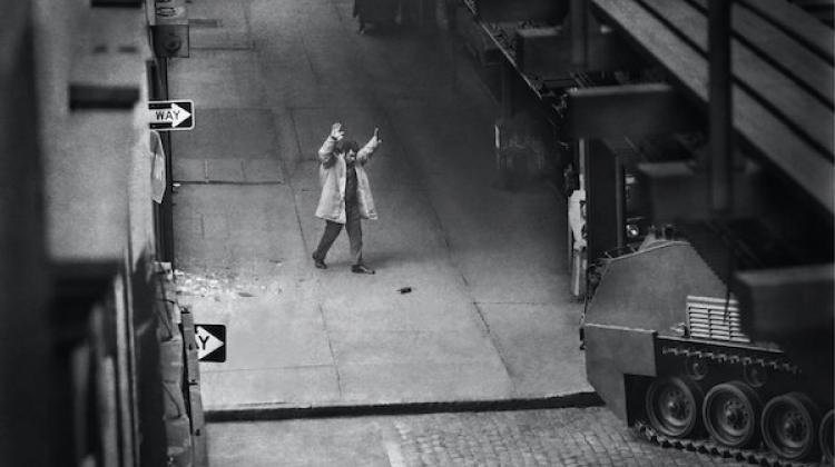 Image shows a man on a sidewalk, with arms raised above his head. In the far right, there appears to be an armored vehicle. The image is in black and white.