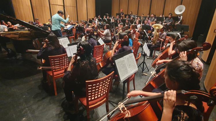 Image of an orchestra, arranged in a small space and facing a conductor and piano