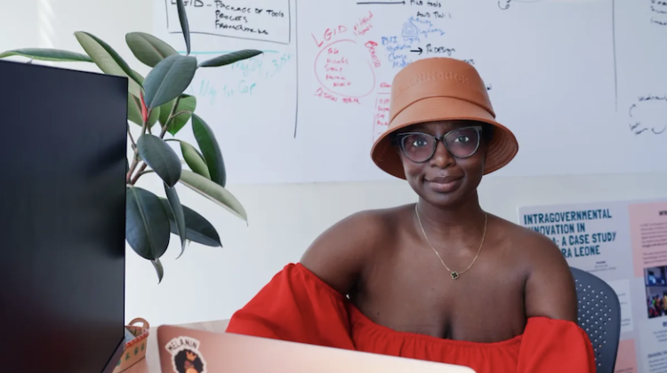 Mariama N'Diaye, in her MIT GOV/LAB office. She wears glasses, a hat, and a bright red top, sitting behind a desk with a laptop open in front of her.