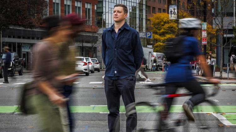 Andres Sevtsuk stands in the middle of a crosswalk as blurry travelers go by.