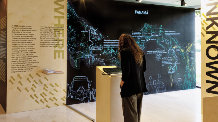 Sarah Williams stands at a podium with her back turned to the camera/viewer, she looks at part of the exhibition Distance Unknown