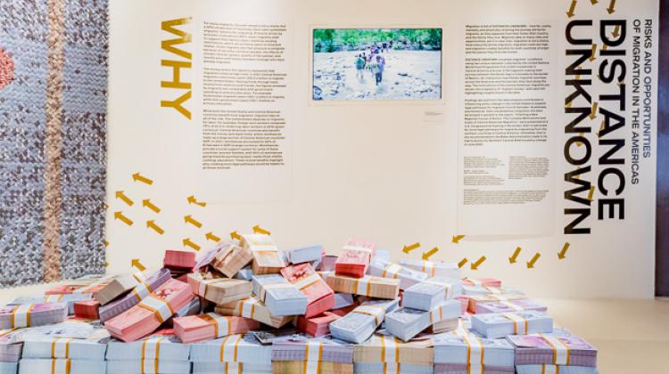 Image of the physical exhibition of Distance Unknown, shoes a pile of designed money in the foreground and exhibition text on a wall in the background.