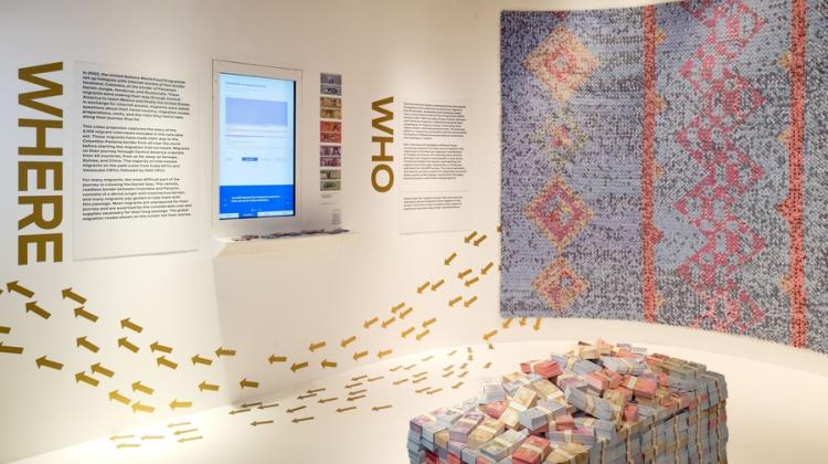 Art exhibit with wall and floor installations prominently features a large stack of bank notes