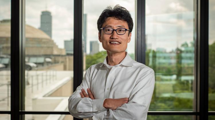 Jinhua Zhao stands with arms folded in front of a window with the Boston skyline in the distance