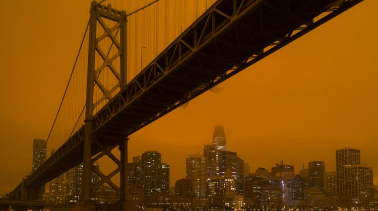Image depicting the orange/red sky produced by wildfires during the summer of 2020. Orientation the shot is under the Bay Bridge, near a support, with the San Francisco Financial District in the background. The spires of the Financial District buildings are cloaked in orange haze.