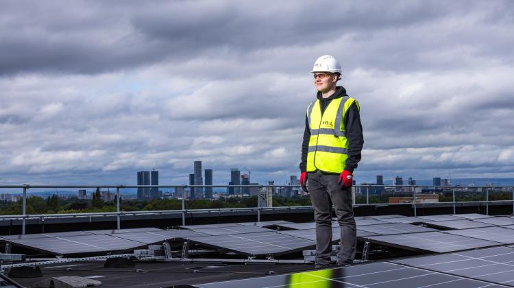 A bank of solar panels with a technician standing over them, in the background one can see a city skyline