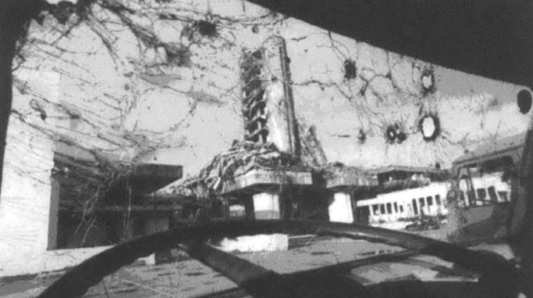 Black and white image taken from inside a SOV, the windshield appears to have bullet holes scattered across its expanse. In the background, ruined buildings can be seen.