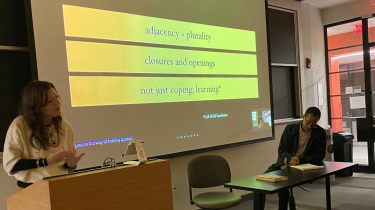 Sara Hendren stands behind a podium and Garnette Cadogan sits at a long table in front of a slide during one of the presentations in this speaker series